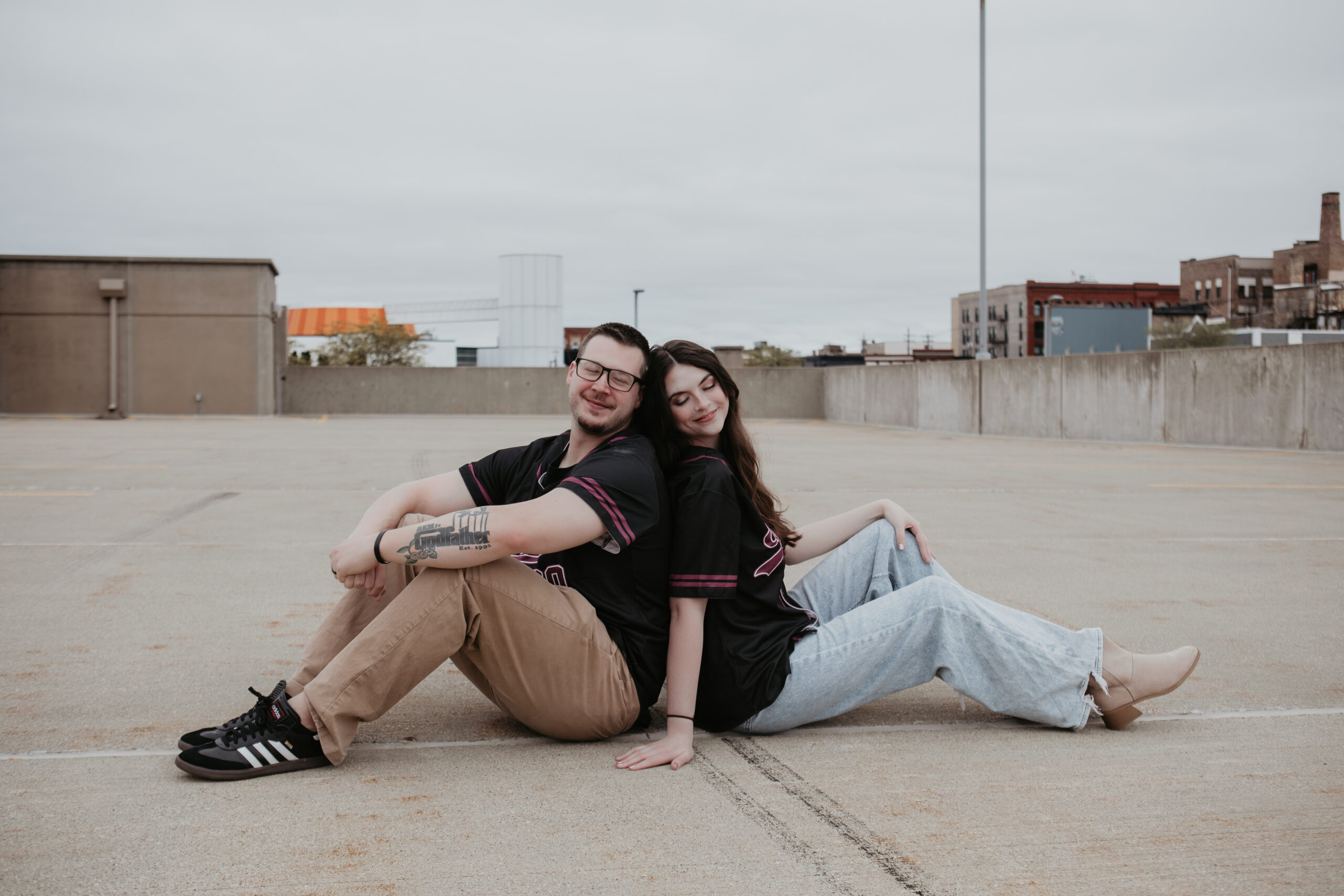 Lansing couple sitting back-to-back during their Downtown Bay City rooftop engagement session, casual and playful vibe.
