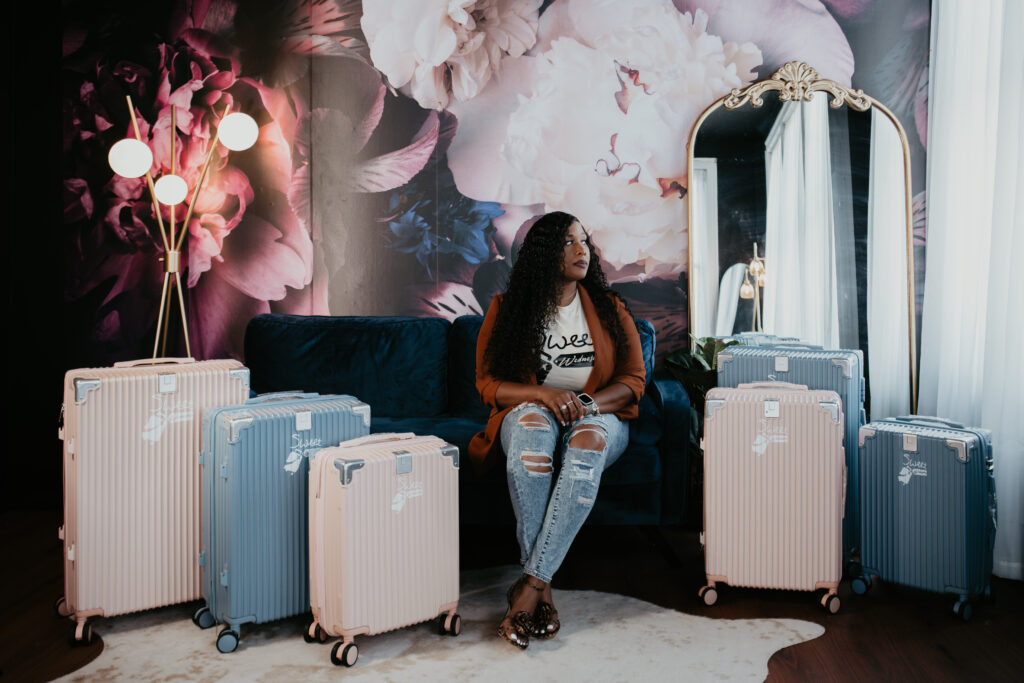 Angela Webb of Sweet Wednesday sits on a navy couch surrounded by pastel blue and blush pink Sweet Wednesday luggage during her branding session in Saginaw, Michigan.