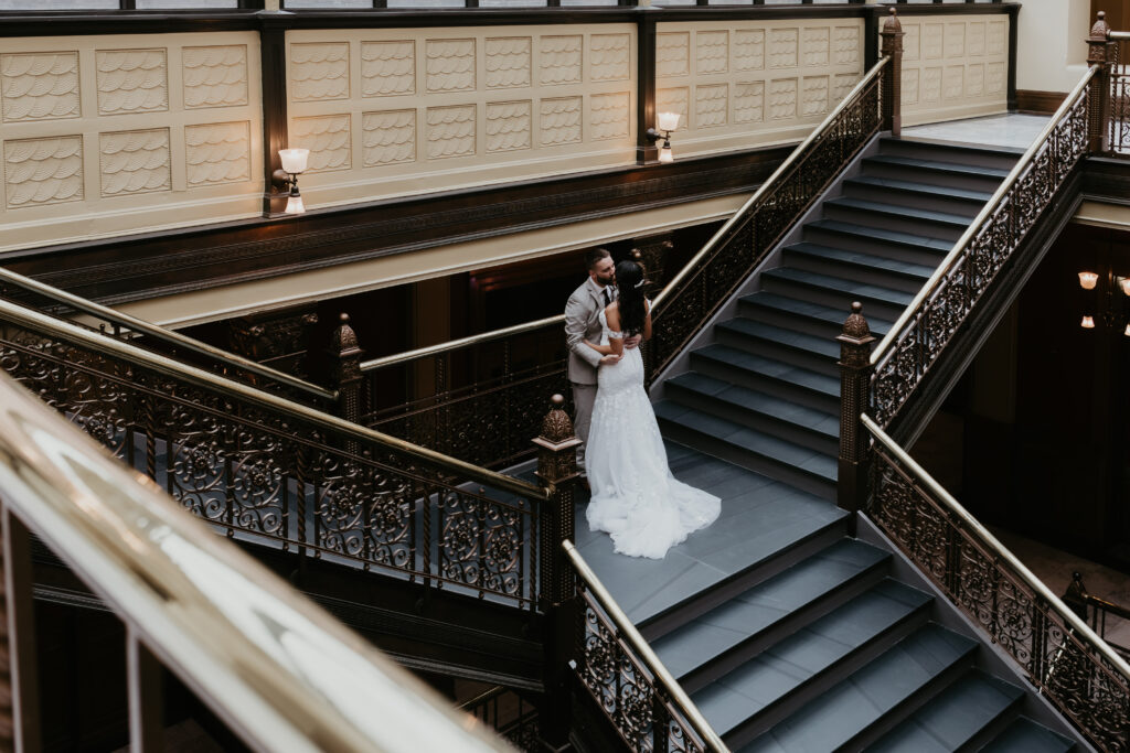 Cassidy and Nate sharing a romantic moment on the staircase inside Bay City City Hall, surrounded by ornate ironwork and warm lighting.