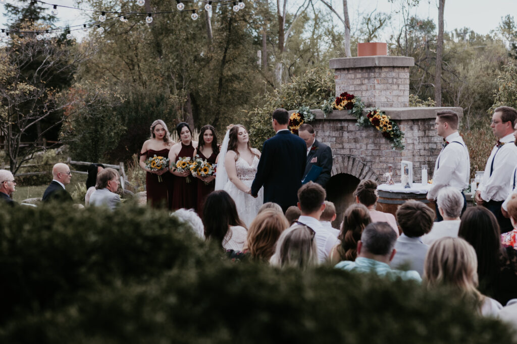 Outdoor wedding ceremony at Fenton Winery & Brewery in Michigan with Nicole and Michael exchanging vows in front of friends and family. Bridesmaids in burgundy hold sunflower bouquets while guests look on.