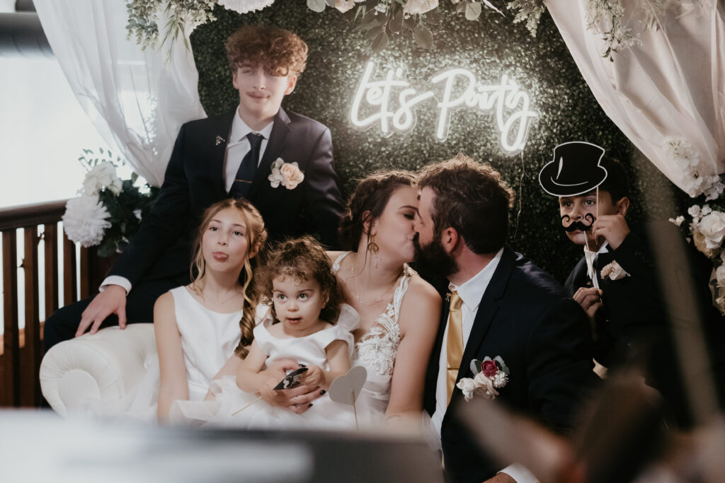 Bride and groom share a kiss surrounded by their kids in a playful wedding photo booth at The Venue at 501 in Clare, Michigan.
