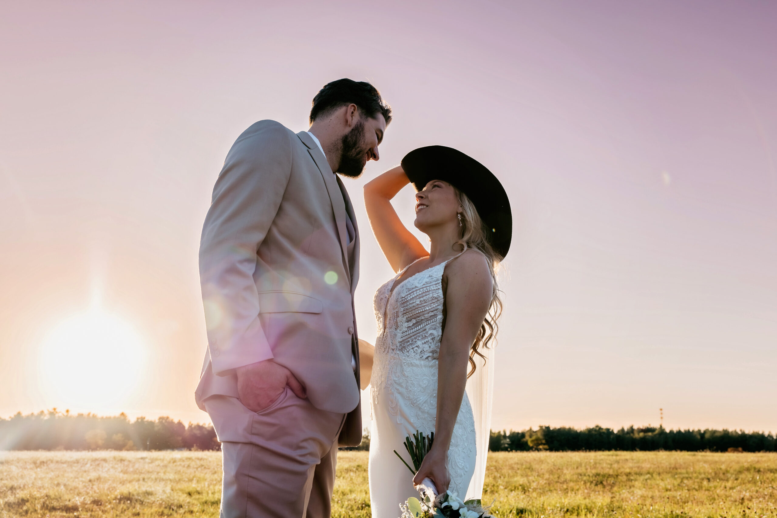 Logan and Nick smile at each other during golden hour in a field at The Barn at Evergreen Farms in Saint Helen, Michigan, wearing western-inspired wedding attire.