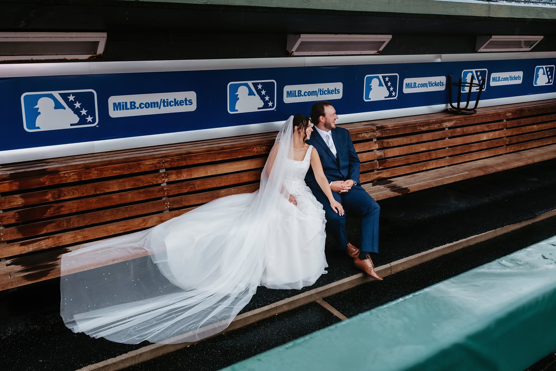 Arissa and Chase sit together holding hands in the baseball dugout at Dow Diamond in Midland, Michigan, with the bride's flowing white wedding gown and veil draped across the wooden bench.